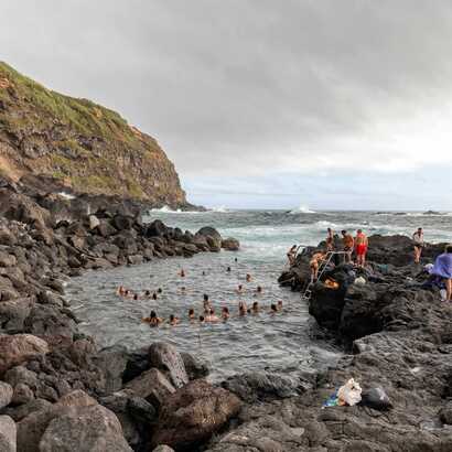 Menschen baden in der natürlichen Meeres-Thermalquelle an der Ponta da Ferraria auf São Miguel, umgeben von Felsen und Atlantikwellen auf den Azoren.