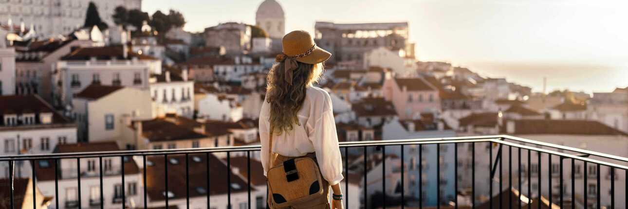 Frau steht auf Balkon und blickt über die Stadt. Die Sonne steht nah am Horizont und taucht alles in ein warmes Licht.