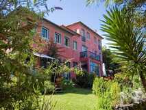 Estrela do Atlantico auf Faial, dreistöckiges, rosarotes Hotelgebäude umgeben von üppigem, grünem Garten mit verschiedenen Pflanzen und Bäumen unter blauem Himmel.
