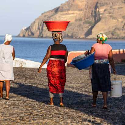 Frauen tragen Behälter in den Händen und auf dem Kopf. Im Hintergrund ist das Meer.