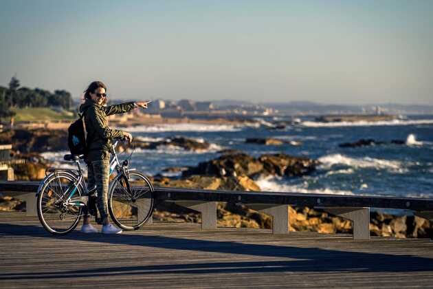 Frau mit Fahrrad steht auf einem Steg und schaut aufs Meer und die Brandung der Atlantikküste Portugals