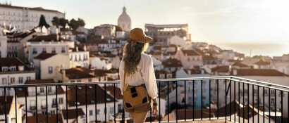 Frau steht auf Balkon und blickt über die Stadt. Die Sonne steht nah am Horizont und taucht alles in ein warmes Licht.