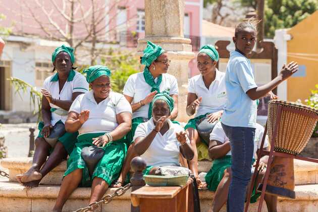 Mehrere Frauen in weiß-grüner Kleidung sitzen draußen zusammen, lachen und klatschen, während ein Junge neben ihnen eine Trommel spielt – typische Alltagsszene auf den Kapverden, die die lebendige, freundliche Straßenkultur zeigt.