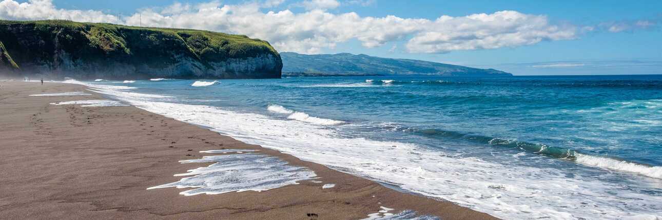 Blick über den schwarzen Sandstrand Praia de Santa Bárbara bei Ribeira Grande auf São Miguel mit Brandung, Steilküste und blauem Himmel.