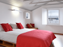 Casa da Baia auf Faial, helles Schlafzimmer mit weißem Holzdeckenbalken, großem Bett mit rotem Bettüberwurf und Kissen, Fenster mit Blick auf den Hafen und die Segelboote.