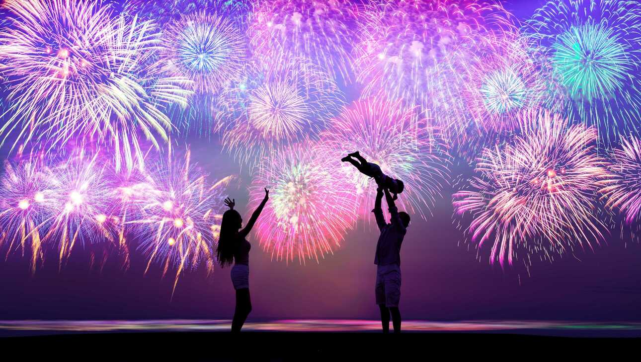 Zwei Menschen feiern mit Wunderkerzen vor einem farbenfrohen Feuerwerk den Jahreswechsel auf den Kapverden.