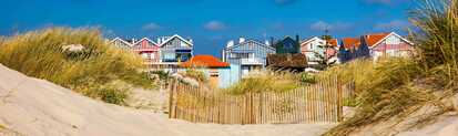 Bunte, gestreifte Strandhäuser stehen hinter sandigen Dünen mit Strandgras, getrennt durch einen hölzernen Dünenzaun, unter blauem Himmel an der Atlantikküste.
