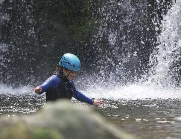 Teilnehmerin mit Kletterhelm genießt das Canyoning-Abenteuer unter einem Wasserfall auf São Miguel.