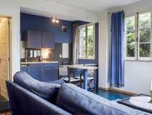 Estrela do Atlantico auf Faial, gemütliches Wohnzimmer mit blauer Couch, kleinen Esstisch und Küchenzeile, helle Fenster mit blauen Vorhängen und natürlichem Licht.