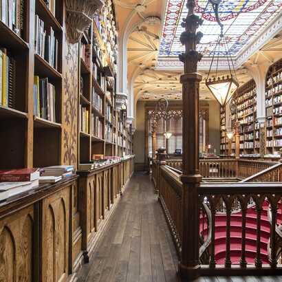 Man sieht einen Gang vorbei verzierter Bücherregale, ein verziertes Glasdach über der roten Treppe, die nach unten führt, in der Livraria Lello, in Porto.