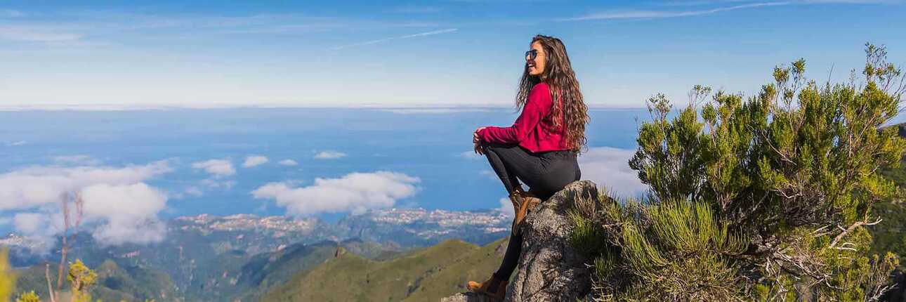 Eine Wanderin steht in wetterfester Kleidung und Wanderschuhen auf dem Gipfel des Pico Ruivo bei klarem Himmel und genießt die Aussicht