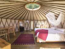 Azul Singular - gemütliches Innenraum-Design einer runden Jurte mit Holzfußboden, Doppelbett mit weißer Bettwäsche und roter Decke, Moskitonetz, Holzmöbeln und natürlichem Licht durch ein rundes Dachfenster.