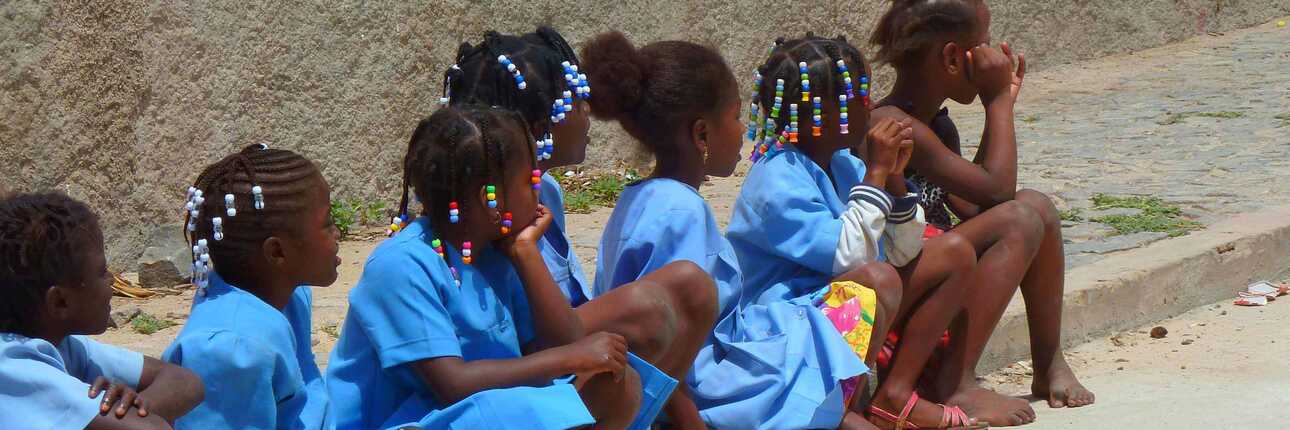 Auf einem Fußweg sitzen mehrere Kinder mit der gleichen Kleidung und bunten Perlen in den Haaren