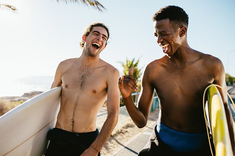 Zwei junge Männer mit Surfbrettern unterhalten sich lachend am Strand der Kapverden – Ausdruck der offenen und herzlichen Sprachkultur.